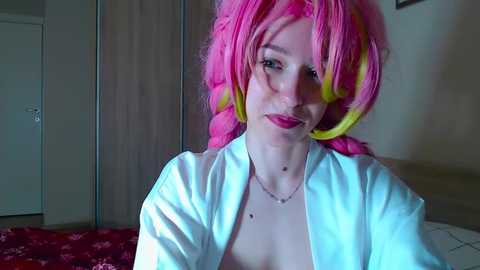Video of a pale-skinned woman with bright pink and yellow hair, wearing a white robe, sitting in a dimly lit bedroom with a red-patterned blanket.