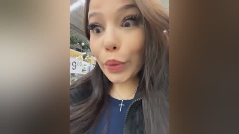 Video of a young woman with long brown hair, fair skin, and dark eyes, wearing a blue top and a black jacket, making a pouty face. Background features a blurred outdoor scene with signs.