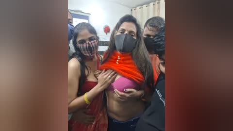 Video of a young woman with a black mask, orange t-shirt, and pink bra, being lifted by two women in red sarees, in a dimly lit room with a curtain and a red flower.