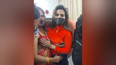 Video of a woman with dark skin and long black hair, wearing a red long-sleeve top, black face mask, and red saree, hugging a woman with long dark hair and a checkered mask. Background shows a beige curtain and another person partially visible.