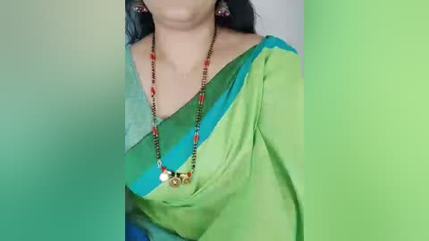A video of a woman in a green and blue sari with a black necklace featuring red and gold beads, wearing a light blue blouse and black earrings. The background is a gradient of green and blue.