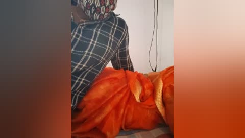 Media: A video of a person wearing a plaid shirt and orange saree sitting on a bed. The background is a plain white wall with a black electrical cord visible. The image is cropped, with a gradient overlay of red and orange.
