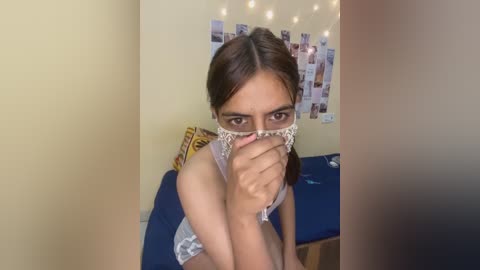 Video of a young woman with medium brown skin and straight brown hair, wearing a white lace bra, standing in a room with yellow walls, blue bed, and string lights.