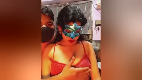 Media: Video of a woman in a red lace bra, blue mask, and black face mask, with a man behind her, in a dimly lit room with a striped curtain.