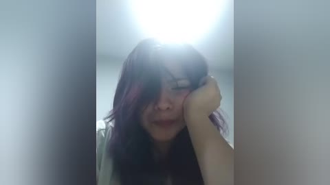 A video captures a young woman with shoulder-length, dark purple hair, resting her head on her hand, with a soft, intimate lighting that highlights her serene expression.