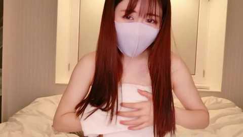 Video of a young Asian woman with long red hair, wearing a white face mask and white towel, seated on a bed with white sheets, in a dimly lit room.