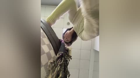 A video of a person in a yellow sweater bending over, revealing a brown headscarf and a patterned skirt, in a tiled bathroom with white walls.