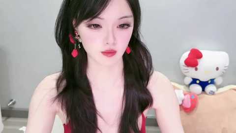 Video of a pale-skinned East Asian woman with long black hair, wearing red lipstick, red earrings, and a red dress, sitting in a room with a Hello Kitty plush toy and a white wall.