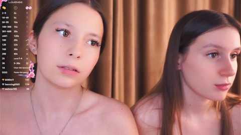 Video of two young women with fair skin, one with long brown hair, the other with shorter hair, both topless, standing side-by-side against a brown curtain background.