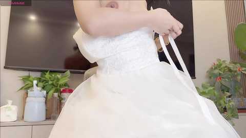 Video of a woman in a white, lace-trimmed strapless wedding gown, holding a white handbag, in a modern living room with a flat-screen TV, potted plants, and decorative items on a white shelf.
