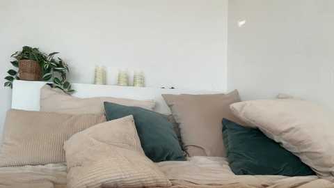 Video of a cozy, minimalistic bedroom with beige bedding, dark green pillows, a white shelf with green plants, and candles, against a white wall.