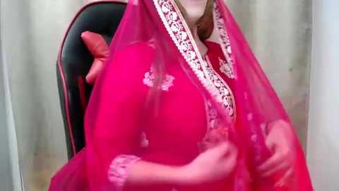 Media: A video shows a woman in a bright pink sari with intricate white embroidery, seated on a black chair. She has light skin and her hands are visible, holding the sari.