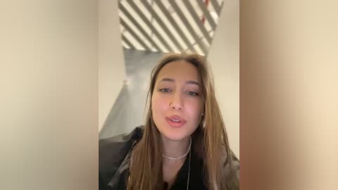 Media: A video of a young woman with long, straight, light brown hair, wearing a black jacket and necklace, standing in a narrow, dimly lit hallway with striped shadows on the walls.