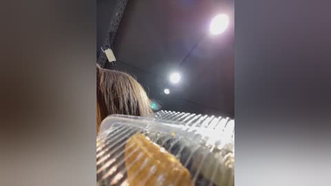 Media: Video of a person with shoulder-length brown hair, partially visible, eating a sticky toffee pudding in a clear glass dish, with a single bright light bulb overhead in a dimly lit, industrial-style room.