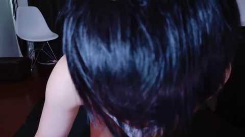 Video of a person with short, dark hair in a sleek, shiny style, captured from a low angle. Background includes a white chair and dark flooring.