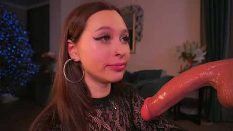 Video of a young, fair-skinned woman with long brown hair, wearing hoop earrings, looking at a large, erect, circumcised penis. She's in a cozy room with a Christmas tree and floral decorations.