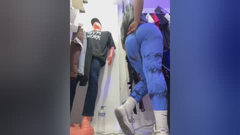 Video of two men in a clothing store. One wears a black T-shirt and blue jeans, standing on an orange box. The other, in tie-dye leggings and white shoes, is in the background, partially visible.