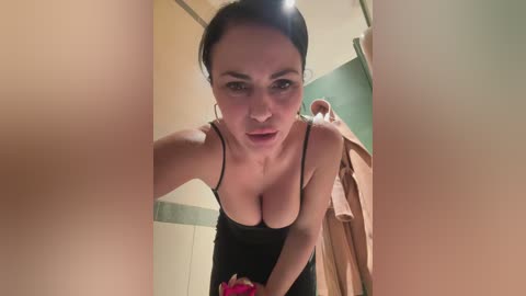 A video of a young woman with light skin, dark hair in a bun, wearing a black camisole, leaning forward with a surprised expression. The background features a green and beige bathroom with a towel hanging.