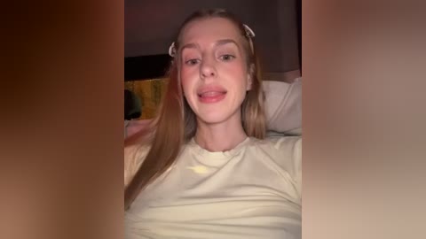 A video of a smiling young woman with light skin, long blonde hair, and wearing a beige t-shirt, lying on a bed with a yellow blanket. The background is dimly lit, creating a cozy, intimate atmosphere.
