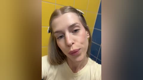 Video of a young woman with light skin and straight, shoulder-length blonde hair, wearing a beige T-shirt, standing in a tiled bathroom with yellow and blue walls.