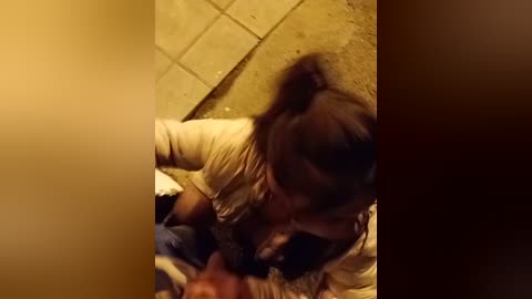 Video of a young person with dark hair, wearing a beige shirt, lying on a tiled floor, kissing a small, brown dog. Warm lighting and shadowy background suggest an indoor setting.