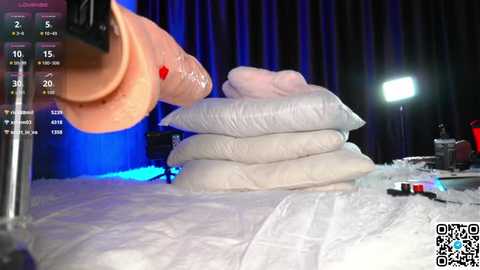 A video of a sex doll with a large, realistic penis being inserted into a pile of white cushions on a bed, with blue and white lighting and a monitor displaying stats on the left.