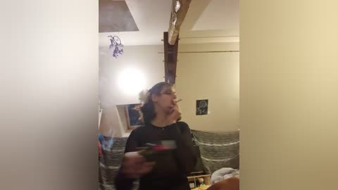 A video captures a woman with curly hair and a dark shirt, holding a smartphone, in a dimly lit room with exposed wooden beams, a gray blanket, and a framed photo on the wall.