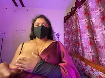 Video of a woman with medium skin tone, black hair, and wearing a black face mask, sitting on a bed with floral curtains in a dimly lit room.