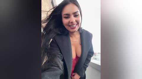 Video of a Latina woman with long black hair, smiling, wearing a black blazer open to reveal a red lace bra, standing indoors near a window.