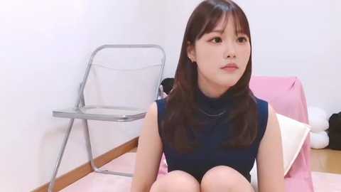 Video of an Asian woman with long black hair, wearing a sleeveless black dress, seated on a pink cushioned chair in a minimalist room with a metal folding chair and wooden floor.