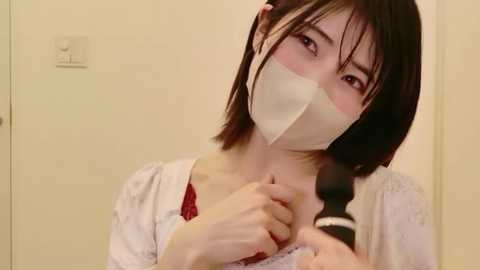 A video of an East Asian woman with shoulder-length black hair, wearing a white mask, a red blouse, and a lace top, holding a black object. The background is a plain beige wall with a small sign.