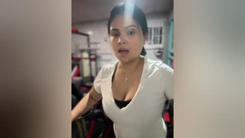 A video of a woman with medium brown skin and dark hair in a ponytail, wearing a white V-neck shirt and black bra, working out in a gym.