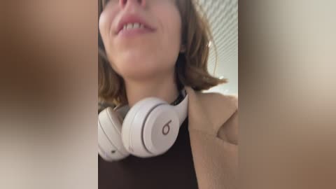 Video of a young woman with light skin and wavy brown hair, smiling, wearing white Beats headphones, and a beige top. Background shows a blurred indoor scene with white blinds.