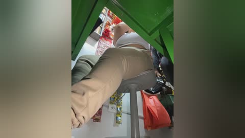 A video taken from a low angle, showing a person's legs in beige pants, climbing into a green container. The background features a cluttered room with colorful objects and a hanging red bag.