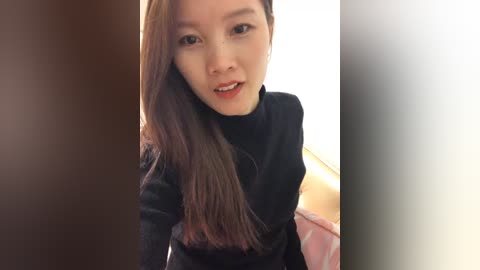 Media: Video of an East Asian woman with long, straight, brown hair, fair skin, and a slight smile, wearing a black turtleneck sweater. The background features a blurred, light-colored room.