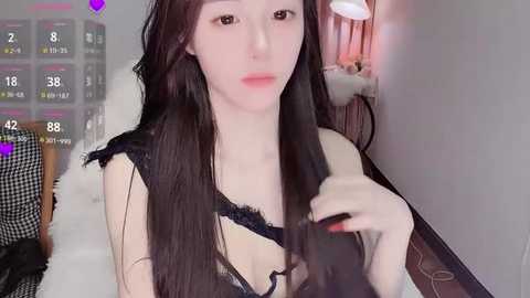 Video of a young Asian woman with long black hair, fair skin, and delicate features, wearing a black lace bra, in a bedroom with a bed and a lamp.