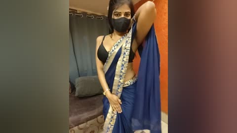 A video of a South Asian woman in a black bra, blue sari, and black face mask, standing in a modern, minimalist room with grey curtains and a brown sofa.