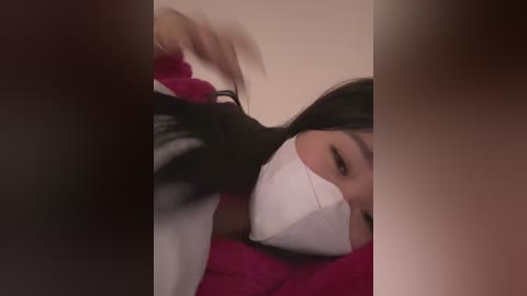 A blurred video showing a woman with long, dark hair, wearing a white face mask, lying on a red blanket. Her eyes are closed, suggesting sleep or rest.