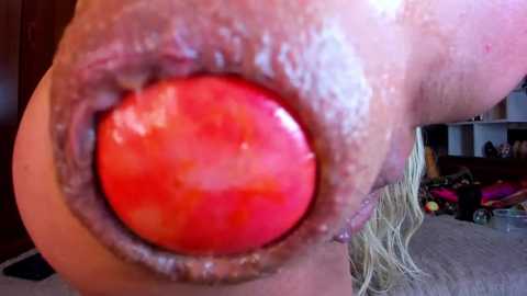 Media: Video of a close-up of a large, shiny, red, heart-shaped nipple, possibly a prosthetic, with a woman's blonde hair visible in the background, and a room with toys in the background.