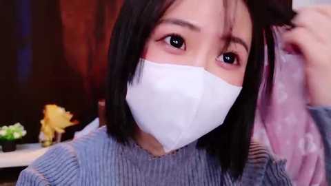 A video of an Asian woman with short black hair, wearing a white surgical mask, adjusting her pink hair tie while indoors, with a blurred background featuring dark and red hues.