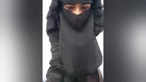 Video of a woman in a black hijab, standing outdoors, wearing a black dress, eyes partially covered by the hijab, and a soft gray gradient background.