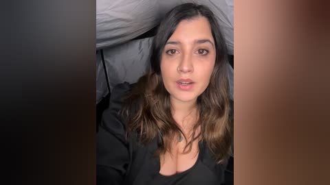 Video of a young woman with medium skin tone and dark brown hair, lying on a bed with grey pillows. She is wearing a black top, and her expression is neutral.