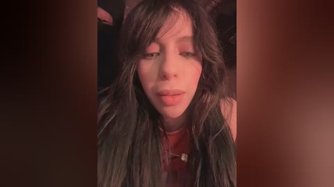 Video of a young woman with long, dark hair, fair skin, and a neutral expression, wearing a red top, partially obscured by a dark background.