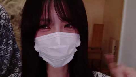 Media: Video of a young Asian woman with long black hair, wearing a white surgical mask, looking concerned. She's indoors, with a patterned background and a blurred figure in the left.