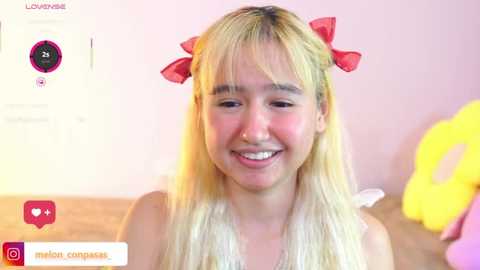 Video of a young, smiling Asian woman with long blonde hair, wearing a red bow headband, in a pink room, holding a yellow plush toy.
