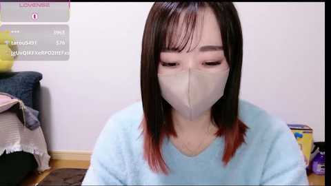 Video of an Asian woman with straight, shoulder-length dark hair, wearing a light blue sweater and a beige face mask, sitting indoors on a wooden floor, surrounded by a cluttered room.
