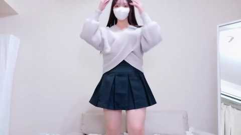Media: Video of an East Asian woman in a white sweater, black pleated skirt, and face mask, adjusting her hair in a minimalist, beige-walled room.