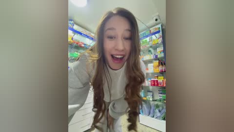 Video of a young woman with long, wavy brown hair, wearing a white sweater, inside a brightly lit pharmacy with colorful shelves full of medicine. She appears surprised and is leaning forward.