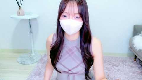 Video of an East Asian woman with long, dark hair, wearing a white face mask, a light pink plaid dress, and sitting on a pink rug in a minimalist room with a white table and a gray chair.