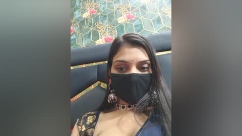 Video of a young woman with long black hair, wearing a black face mask, blue denim jacket, and a black lace top, seated on a black upholstered chair with colorful hexagonal patterned wallpaper in the background.